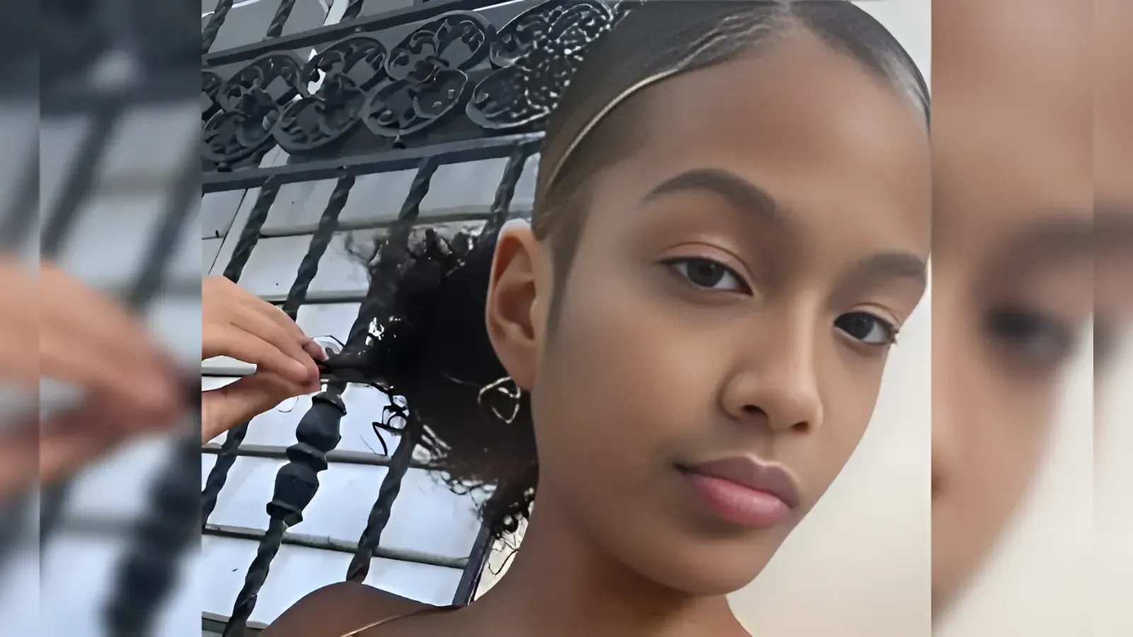Close-up portrait of a young woman with sleek braided hair while a stylist braids the back, iron railing in the background