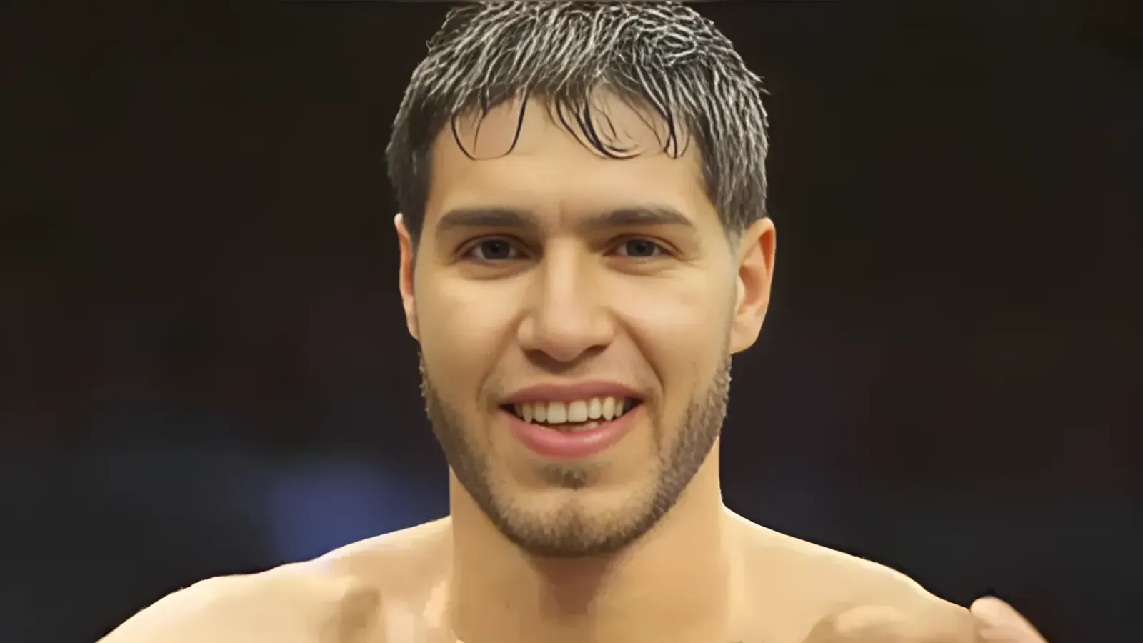 Close-up of a smiling man with short dark hair and stubble, shirtless against a dark background.