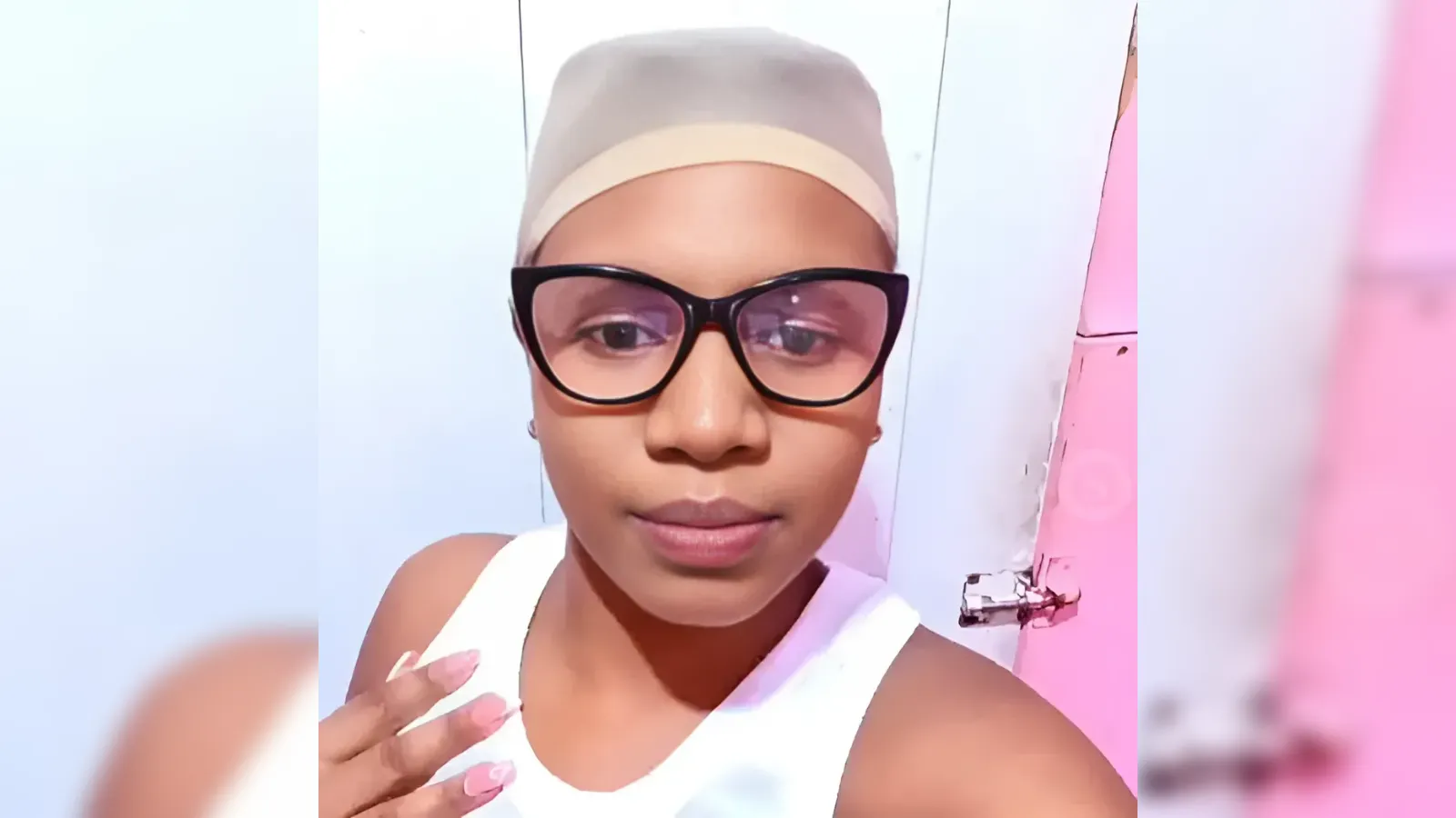 Young woman wearing a beige head wrap and black-framed glasses, taking a selfie with a pastel pink background behind her