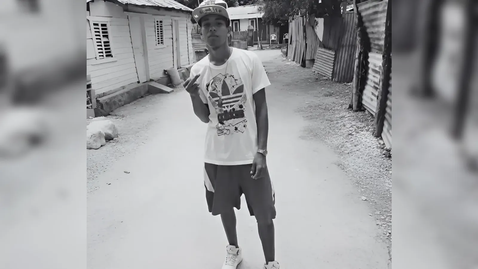 Young man in a cap and graphic T-shirt stands in a dirt street, making a rock-on hand gesture in a black-and-white street scene.