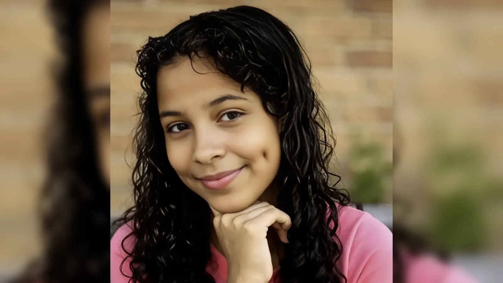Young woman with dark curly hair in a pink top, resting chin on her hand and smiling at the camera in a blurred outdoor background.