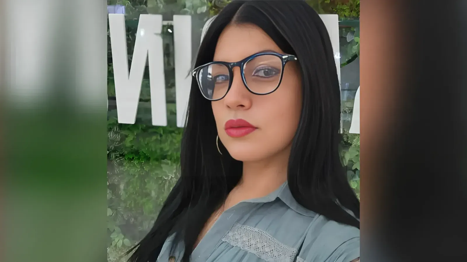 Portrait of a woman with long dark hair and blue-framed glasses outdoors, wearing a light blue shirt with lace detail.