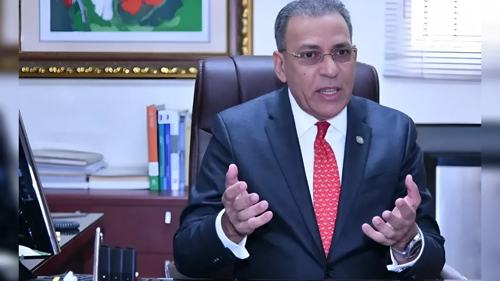 Man in a dark suit and red tie speaks while gesturing with hands in an office setting.