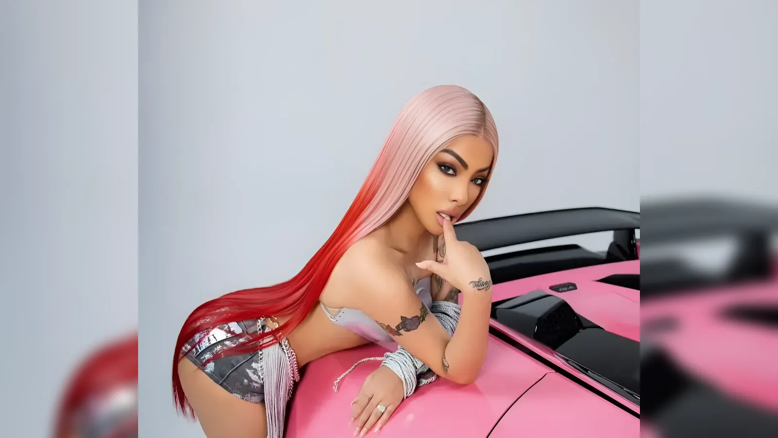Woman with long pink-to-red gradient hair posing on a pink sports car, looking at the camera with a finger to her lips.