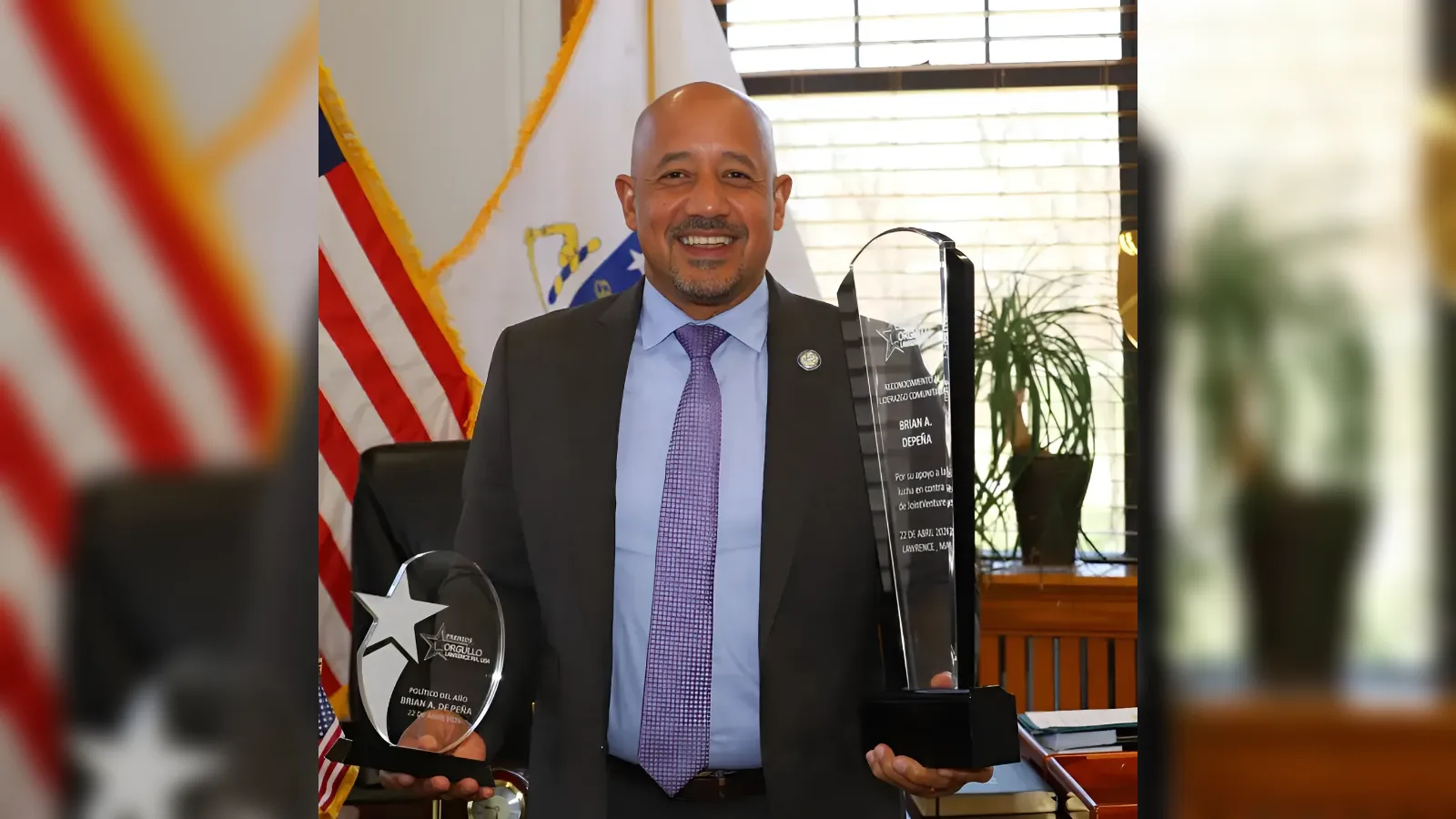 Smiling man in a suit holds two glass trophies in an office with an American flag in the background.