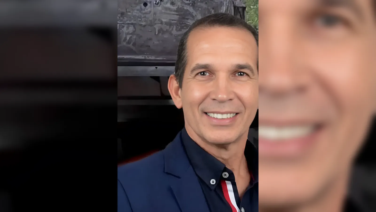 Close-up selfie of a smiling man in a dark navy blazer and navy shirt, posing for the camera outdoors.