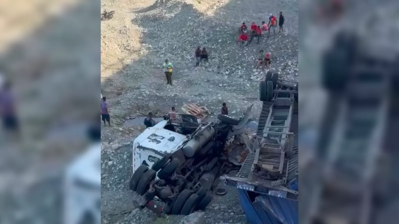 Overturned large truck on a rocky slope with its cab crushed; bystanders and responders stand nearby inspecting the scene.