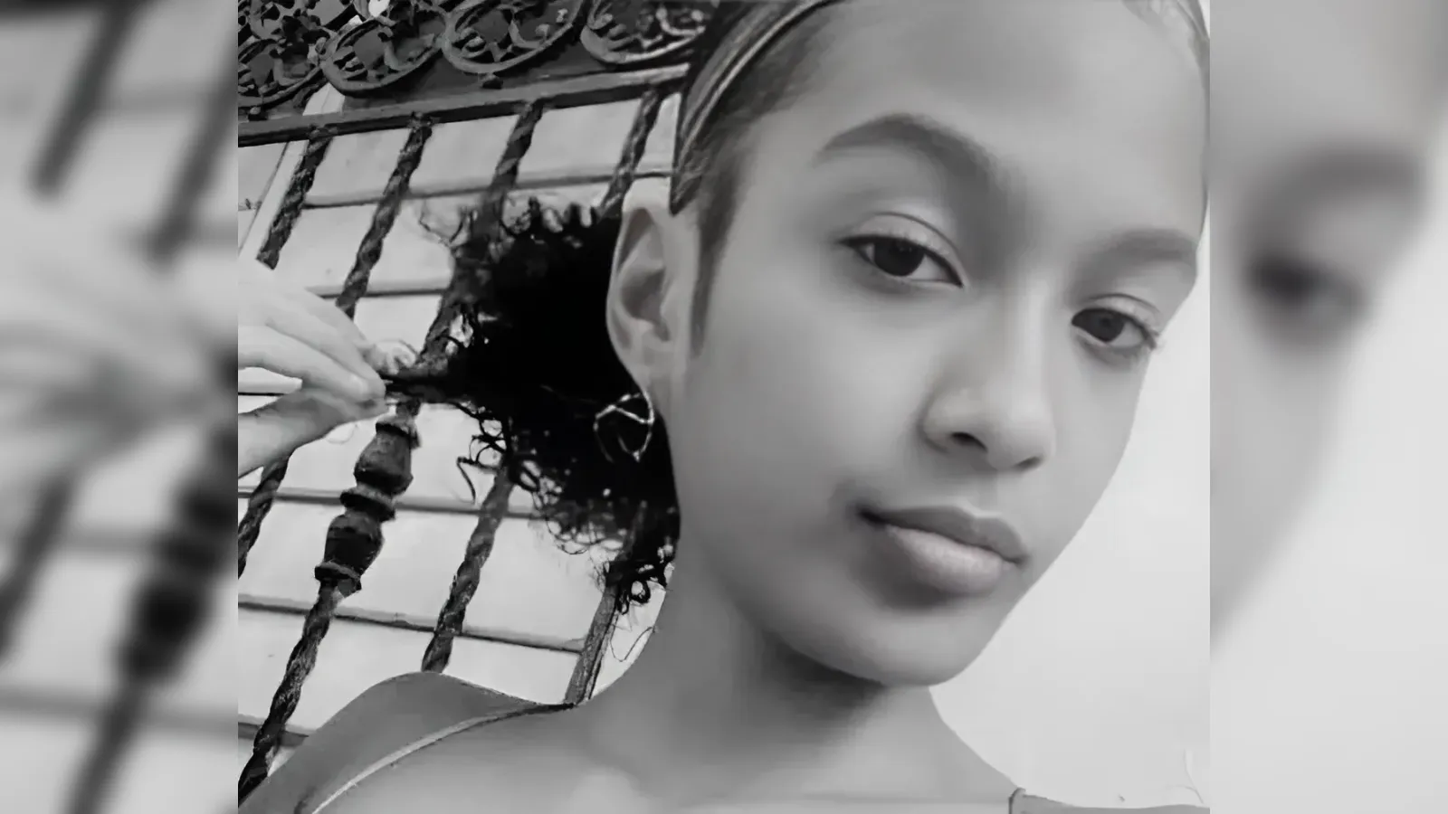 Close-up black-and-white portrait of a young woman looking at the camera, touching her curly hair by a decorative fence nearby.