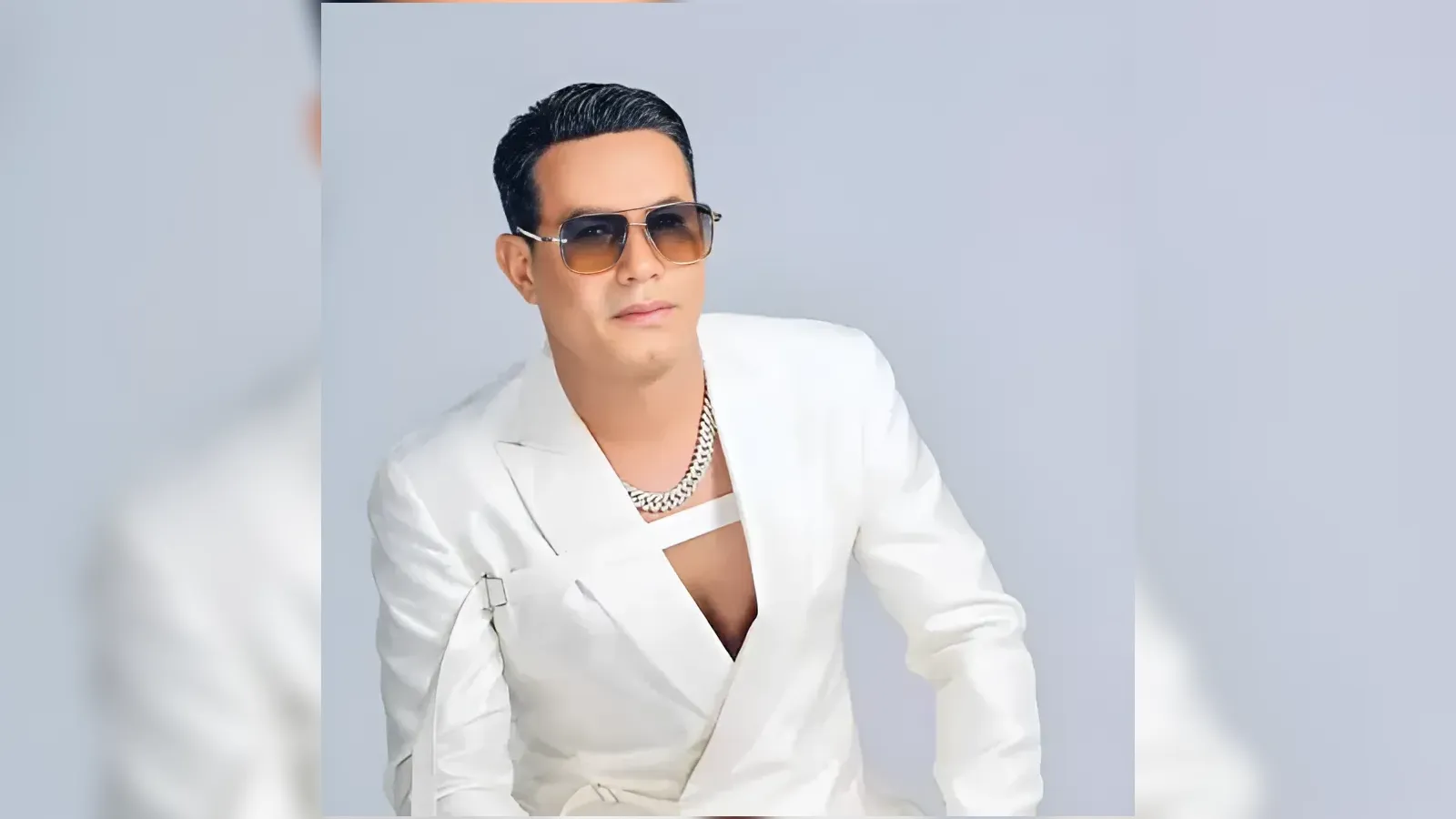 Man in a white blazer with a chunky chain necklace and sunglasses posing against a pale blue background.
