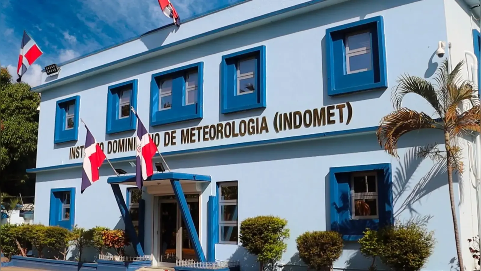 Facade of the Instituto Dominico de Meteorologia (INDOMET) building, light blue with blue-framed windows and Dominican flags outside