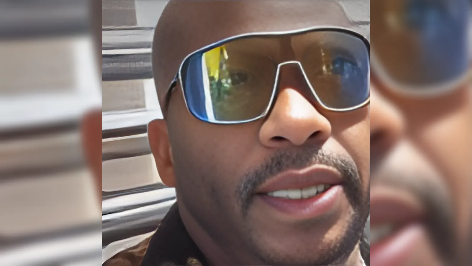 Close-up of a man with a shaved head wearing large mirrored sunglasses, smiling slightly inside a car.