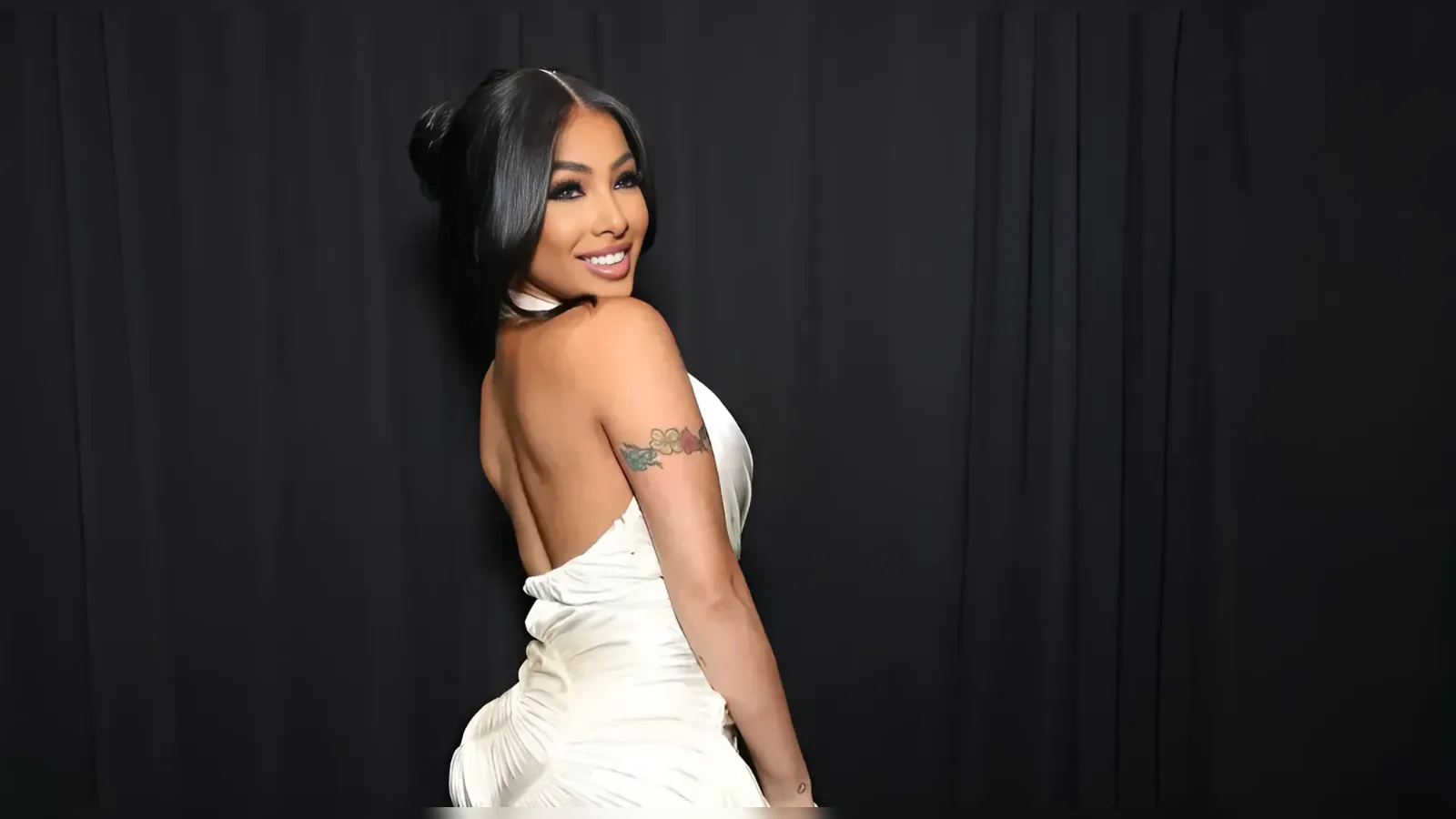 Smiling woman in a white backless gown posing sideways against a dark backdrop, with a flower tattoo on her upper arm.
