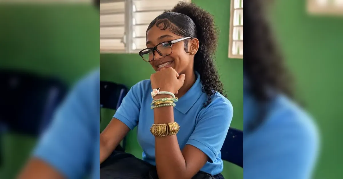 Smiling girl with braces wearing a blue polo and many bracelets, sitting in a green-walled classroom with a window behind her.