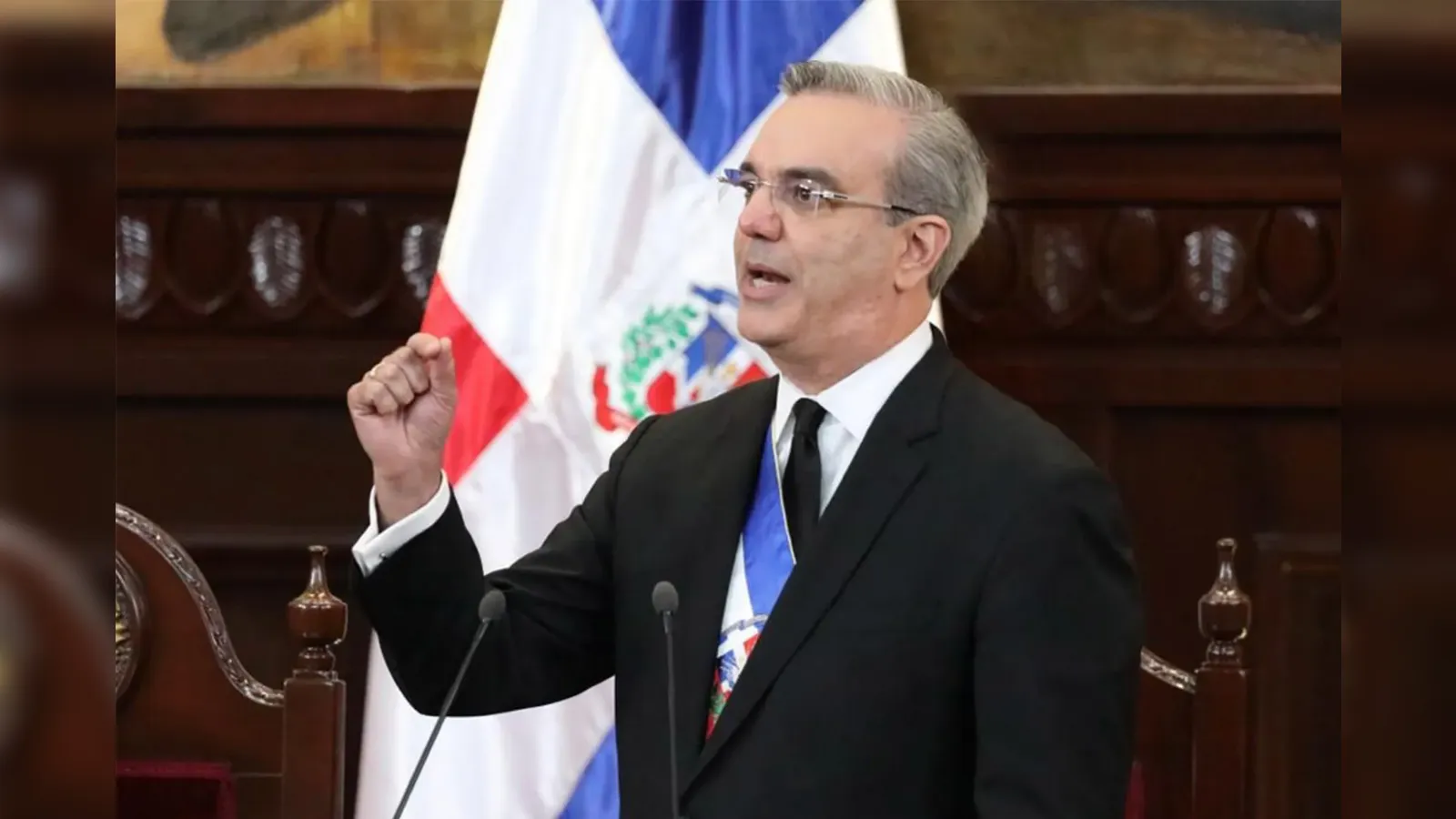 Man in a black suit giving a formal speech at a podium, with a flag in the background and microphones nearby.