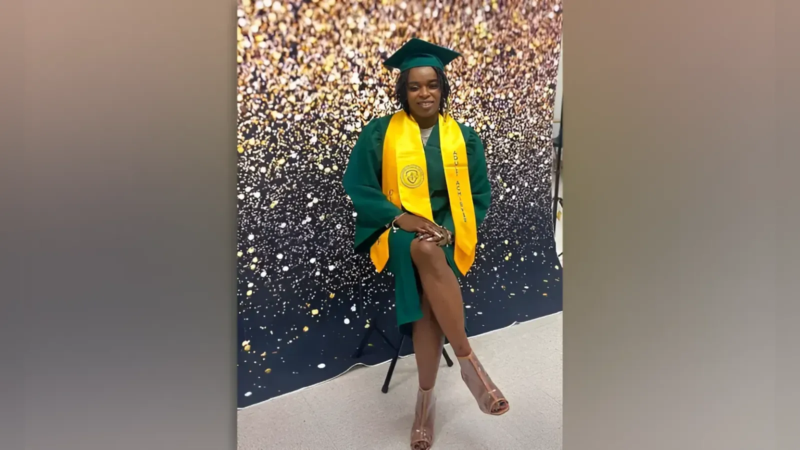 Graduate in a green cap and gown with a yellow stole, posing in front of a glittery backdrop.