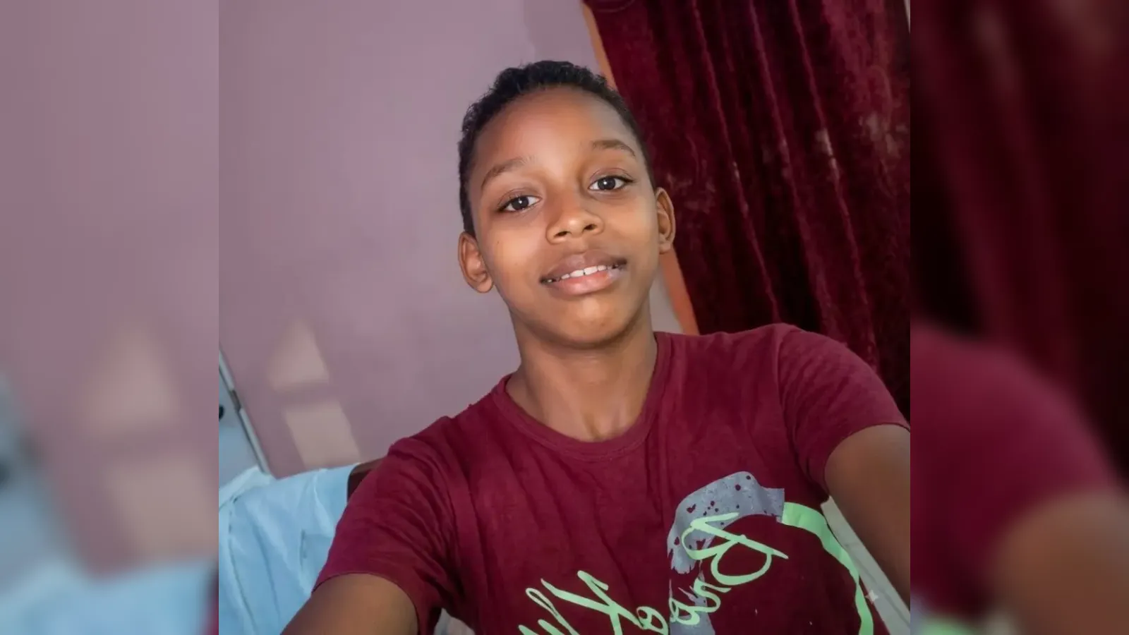 Young person taking a selfie indoors, wearing a maroon shirt, with a purple wall and burgundy curtain in the background.