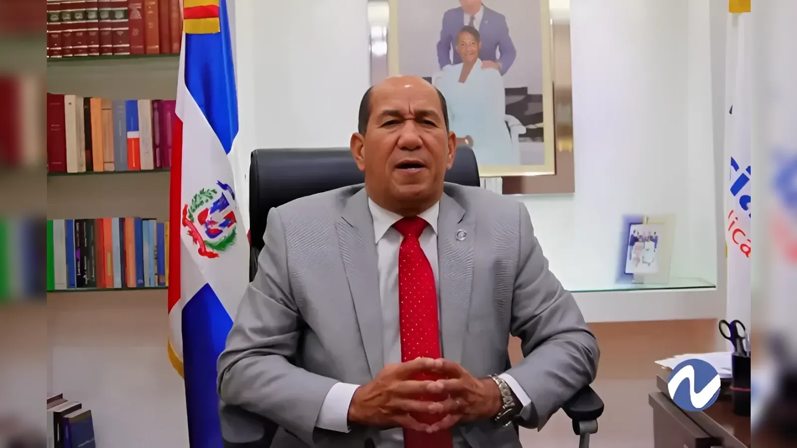 Man in a grey suit and red tie sits at a desk in an office, hands clasped, with the Dominican flag behind him and a framed photo on the wall behind.