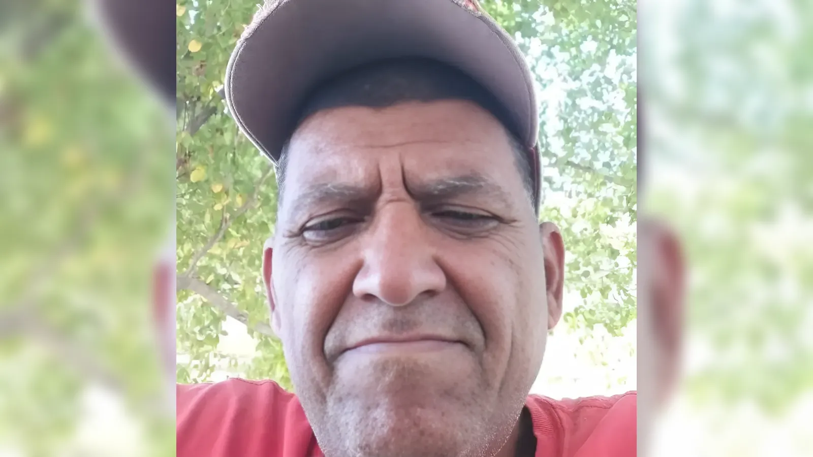 Close-up selfie of a man wearing a brown cap outdoors, with a neutral expression and green trees in the background, wearing a red shirt.”,