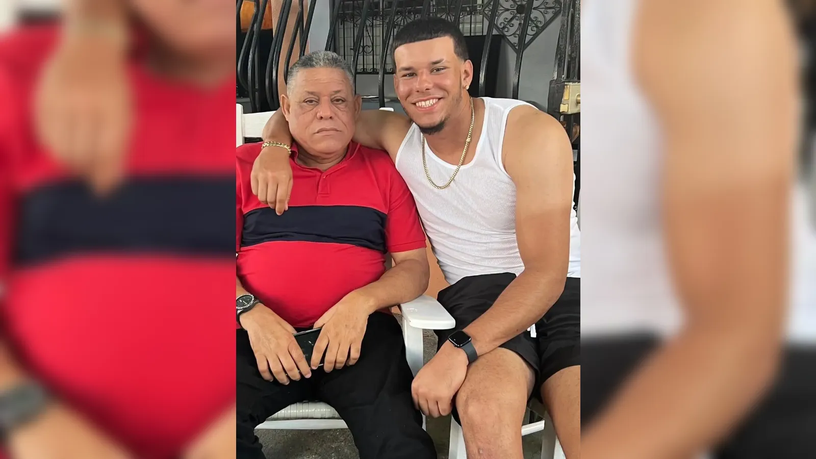 Two men sit close together on outdoor chairs; older man in a red polo, younger man in a white tank top with a gold chain, smiling at the camera.