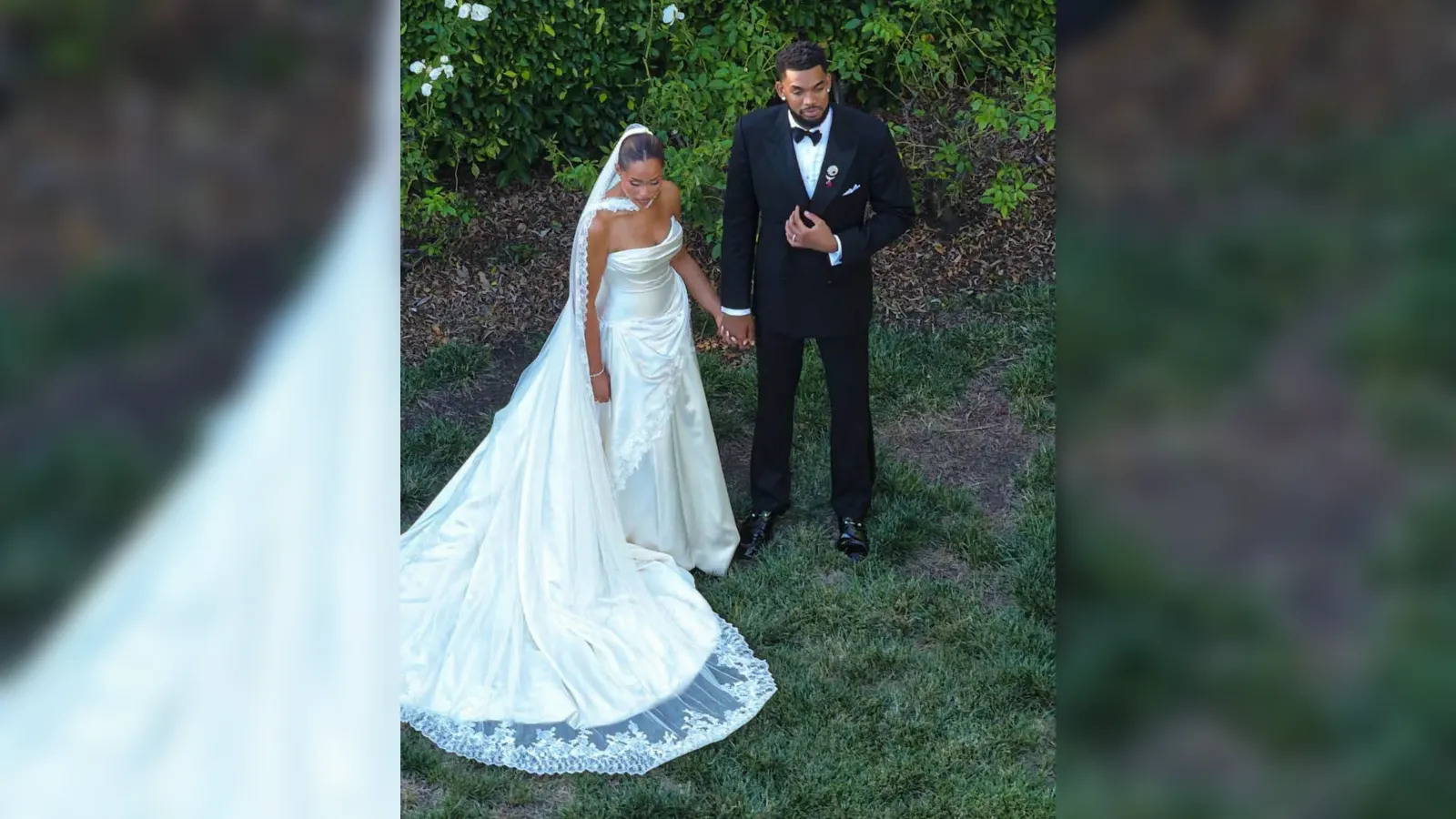Bride in a white wedding gown with long train and veil holds hands with a groom in a black tuxedo in a garden setting.