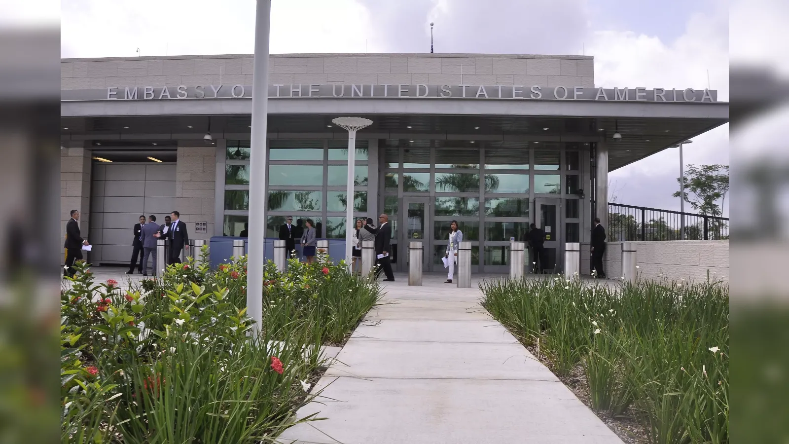 People in business attire stand outside the U.S. Embassy entrance on a walkway with plants nearby and glass doors behind them.