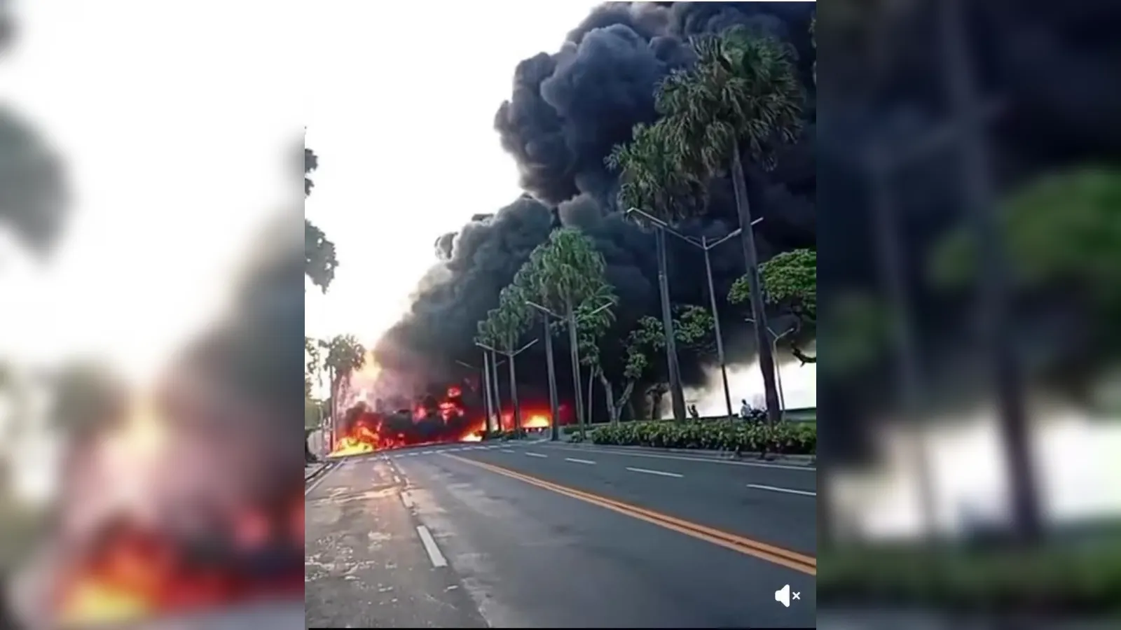 Vehicle fire on a wide road with tall black smoke and flames, palm trees lining the street and emergency activity visible in the distance.
