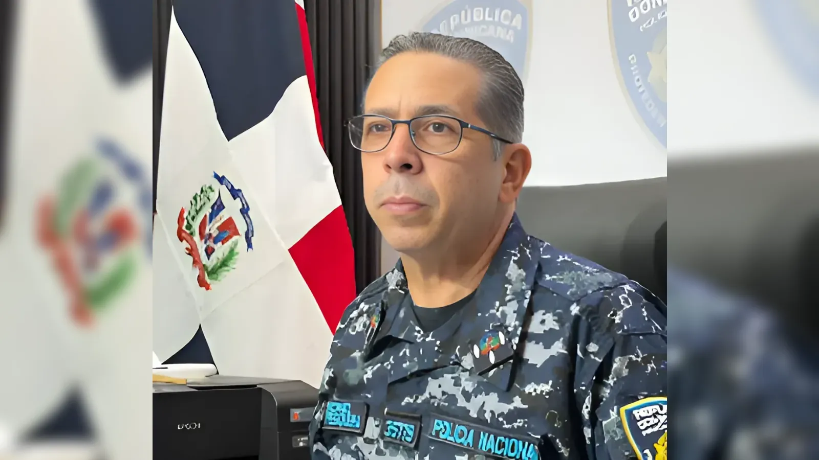 Portrait of a Dominican military or police officer in camouflage uniform, wearing glasses, seated at a desk with the national flag in the background.