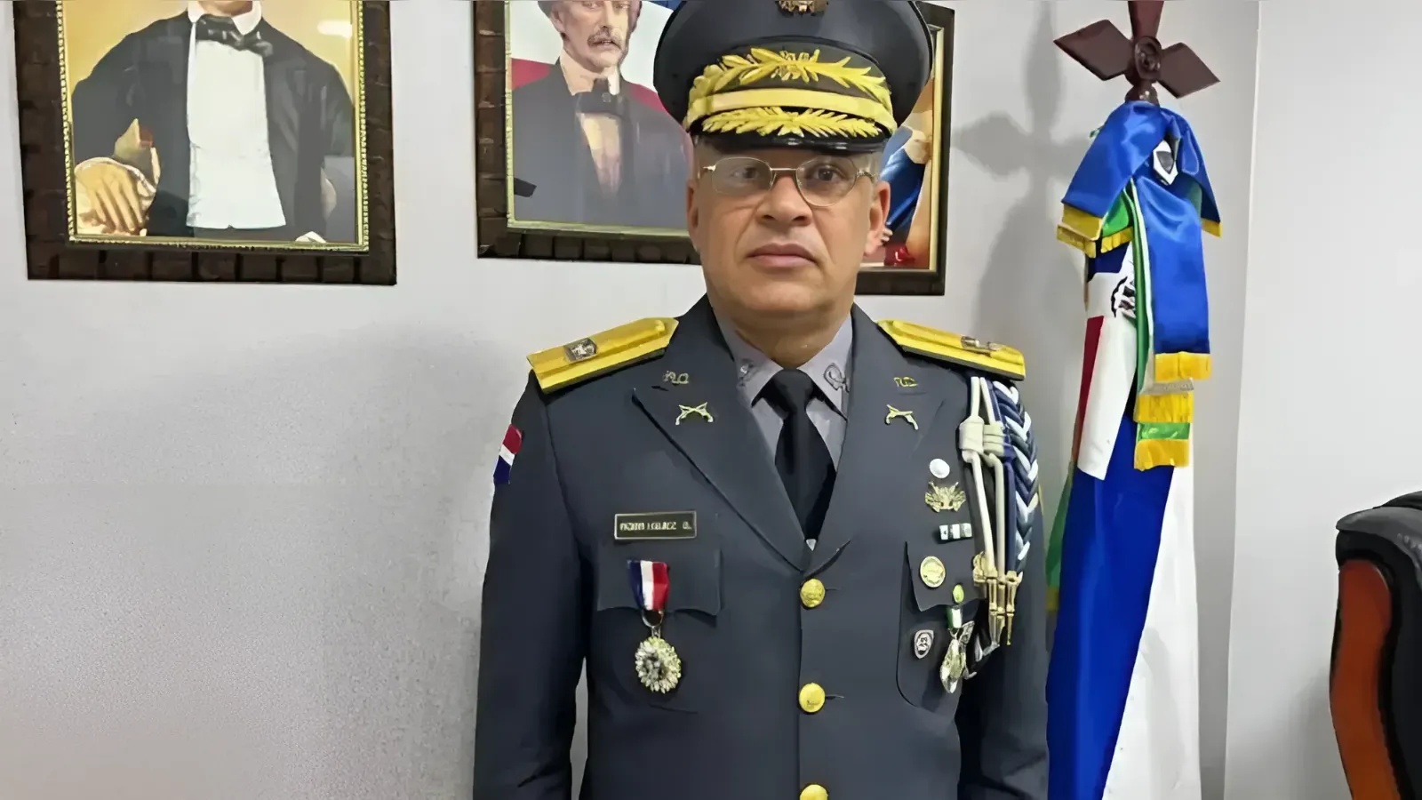 Formal portrait of a male military officer in dress uniform with gold epaulettes and medals, standing indoors with framed portraits and a flag in the background