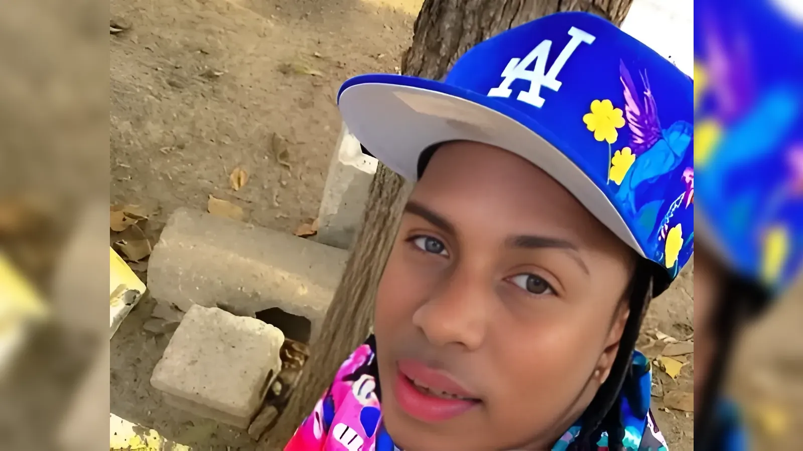 Close-up of a young woman wearing a blue LA cap with yellow flowers, outdoors beside a tree.
