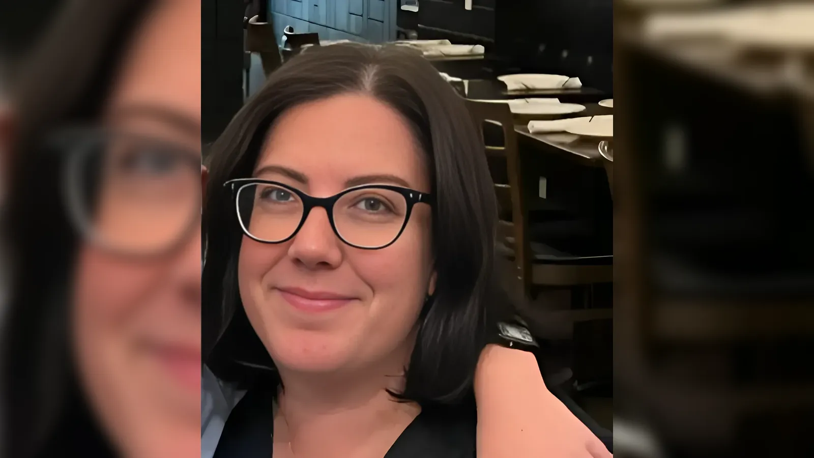 Smiling woman with short dark hair and black glasses in a casual indoor restaurant setting, close-up portrait.