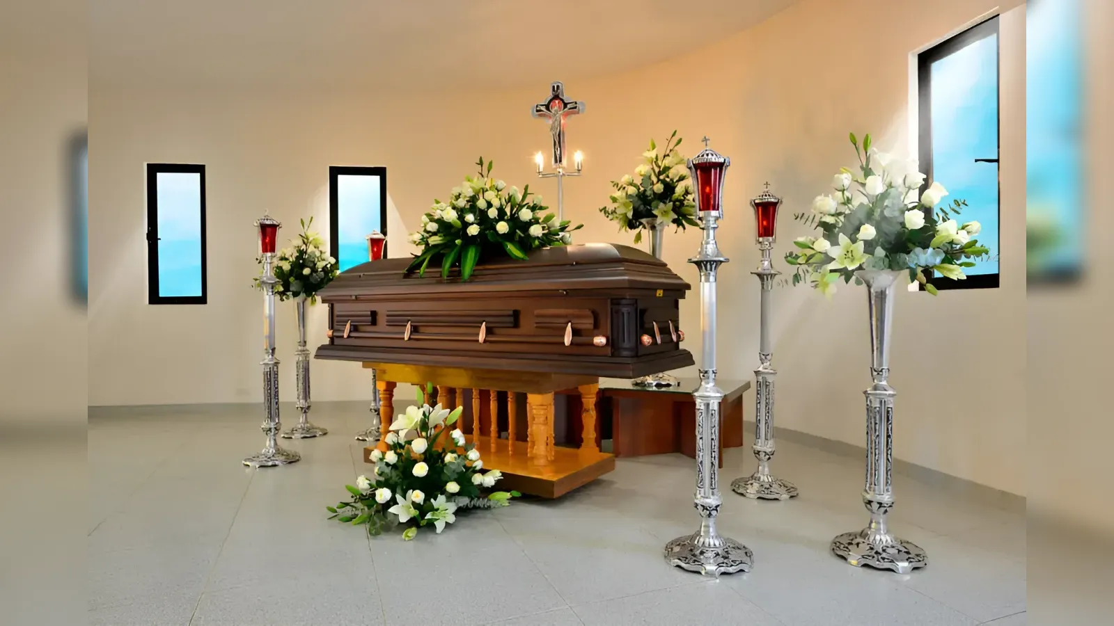 Wooden funeral casket on a stand, surrounded by white flowers and tall silver candle holders in a chapel setting.