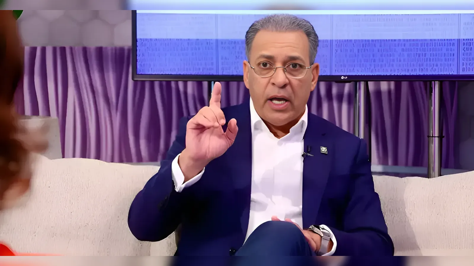 A man in a navy suit gestures with his index finger raised while speaking on a talk-show set.