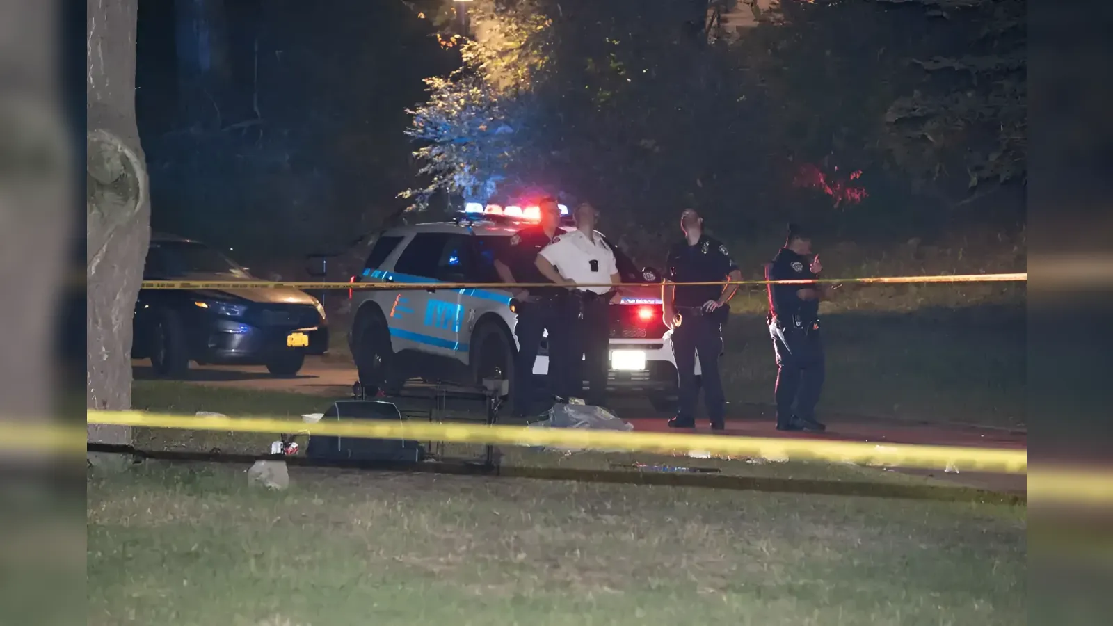 Nighttime police scene with officers behind yellow caution tape, a patrol SUV with flashing lights, and scattered debris on the ground.