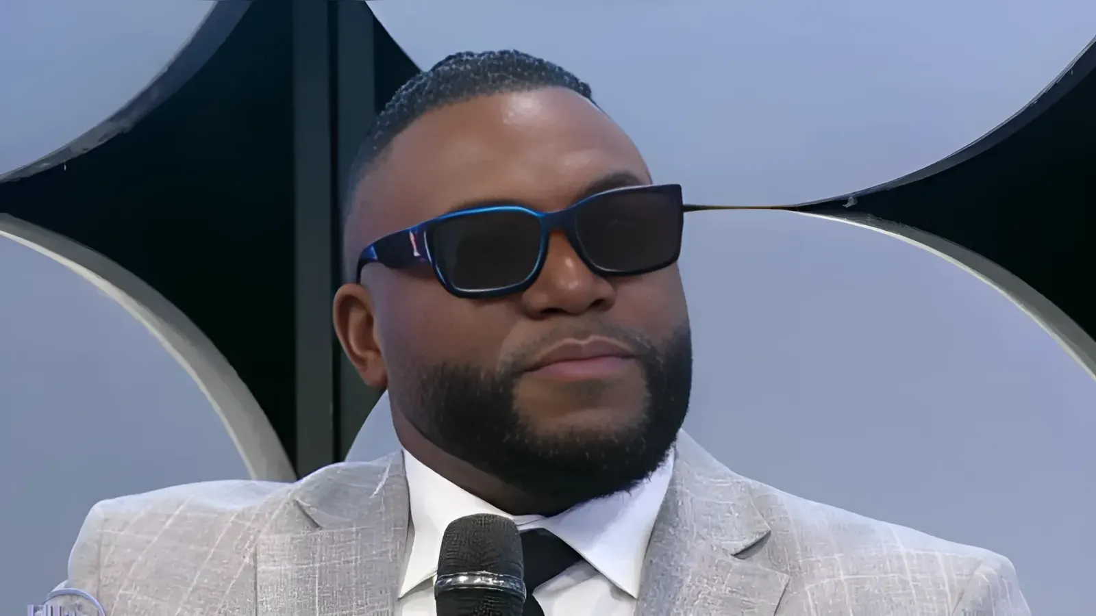 Close-up of a man in a light gray suit and white shirt wearing dark sunglasses, speaking into a handheld microphone on stage.