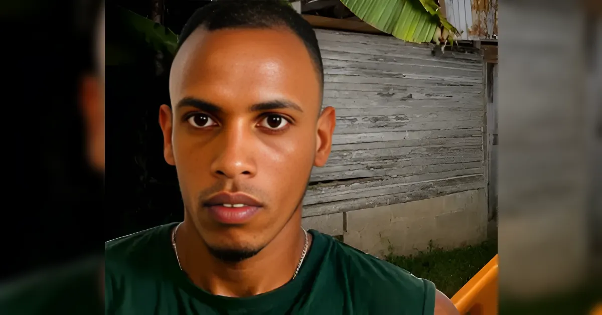 Close-up of a young man with short hair and a green sleeveless shirt, outdoors at night beside a weathered wooden wall.