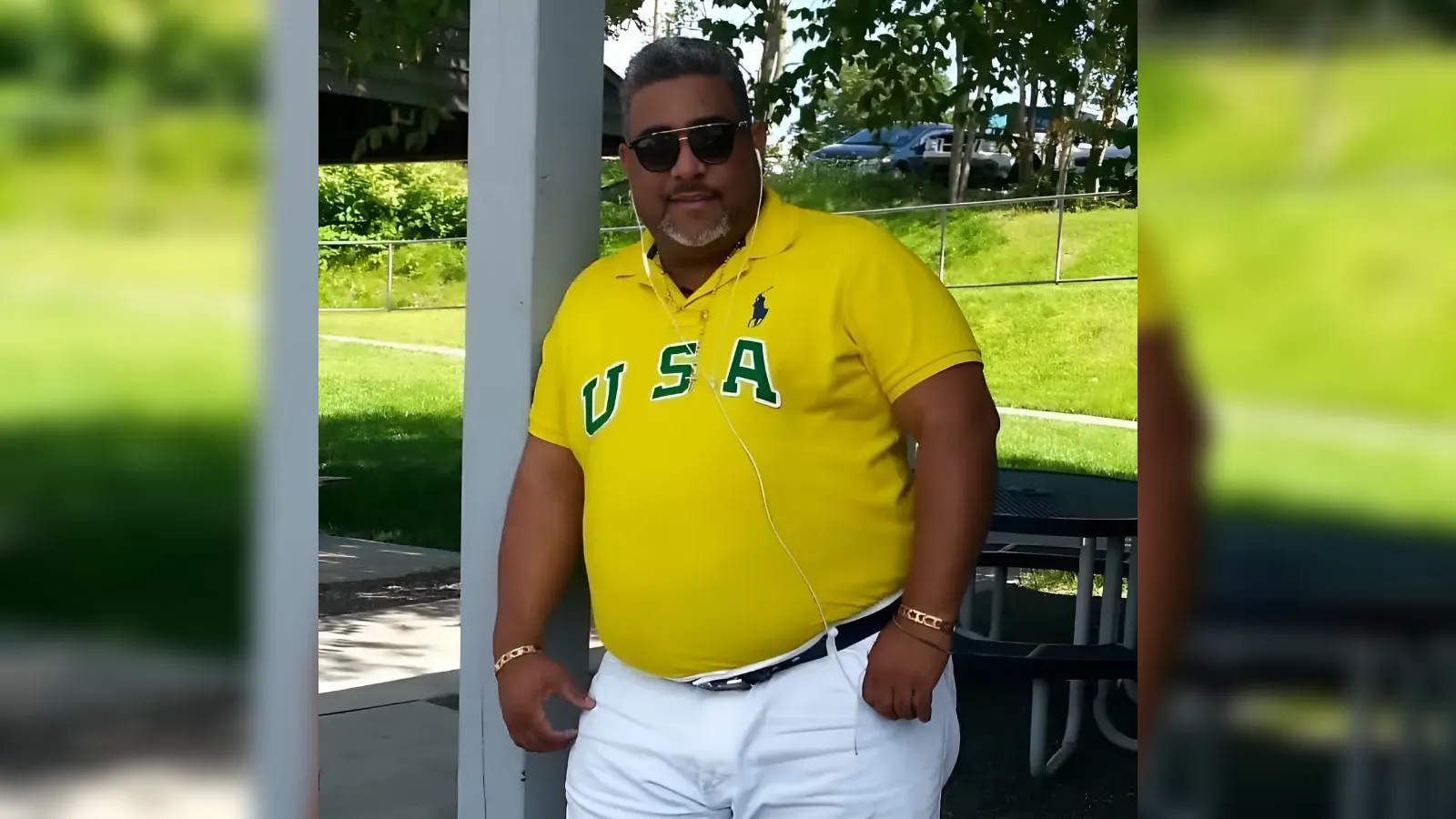 Man in a bright yellow USA polo and white shorts stands outdoors wearing sunglasses and earphones, under a shaded area.