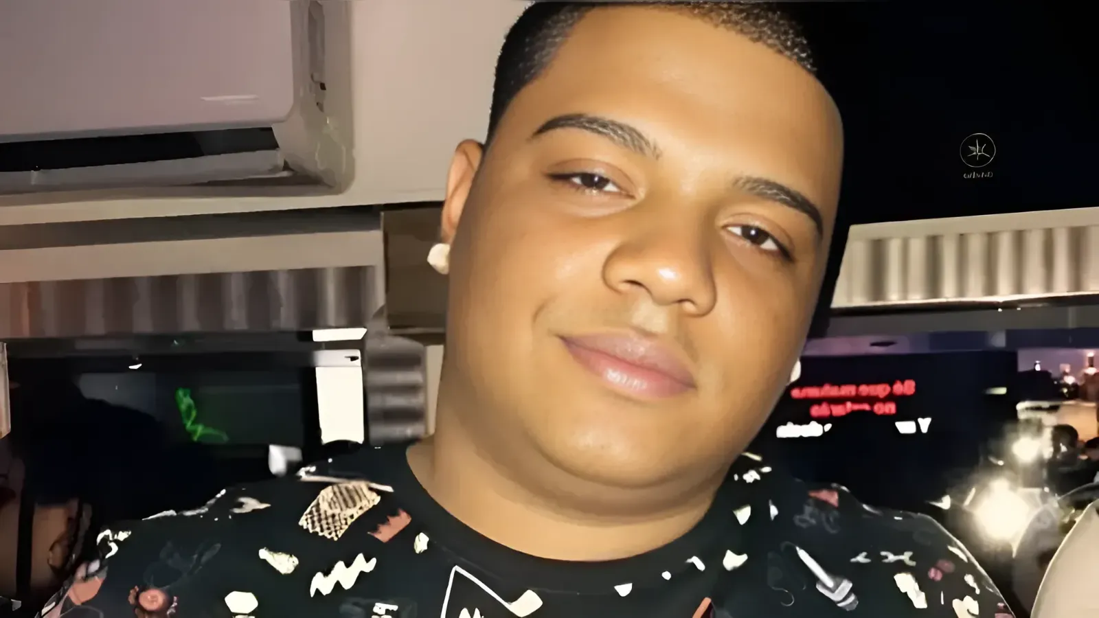 Close-up portrait of a smiling young man wearing a patterned black shirt, indoors at night.