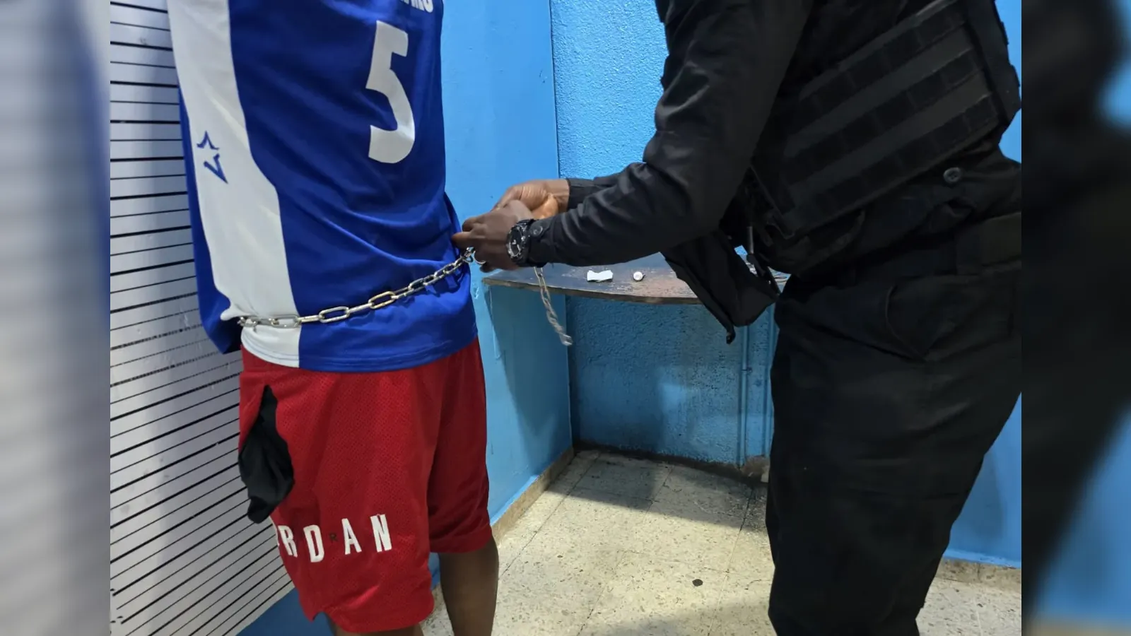 Person in a blue and white jersey and red shorts is being handcuffed by a uniformed officer in a black outfit beside a blue wall.