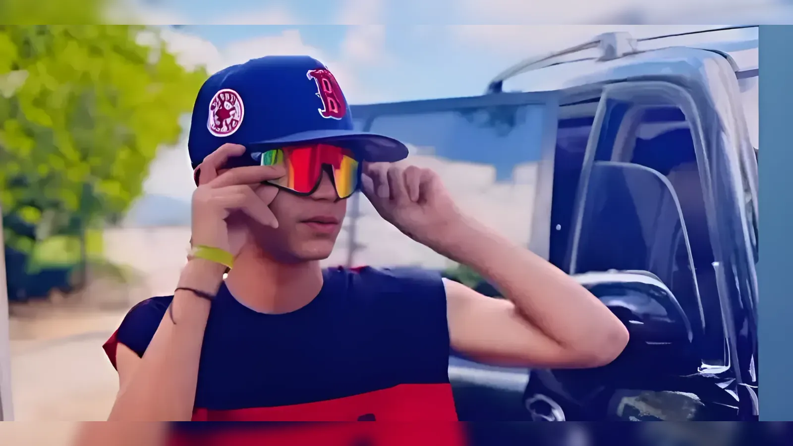 Young man wearing a blue cap with a red logo and rainbow mirrored sunglasses, adjusting them beside an open car door on a sunny day.