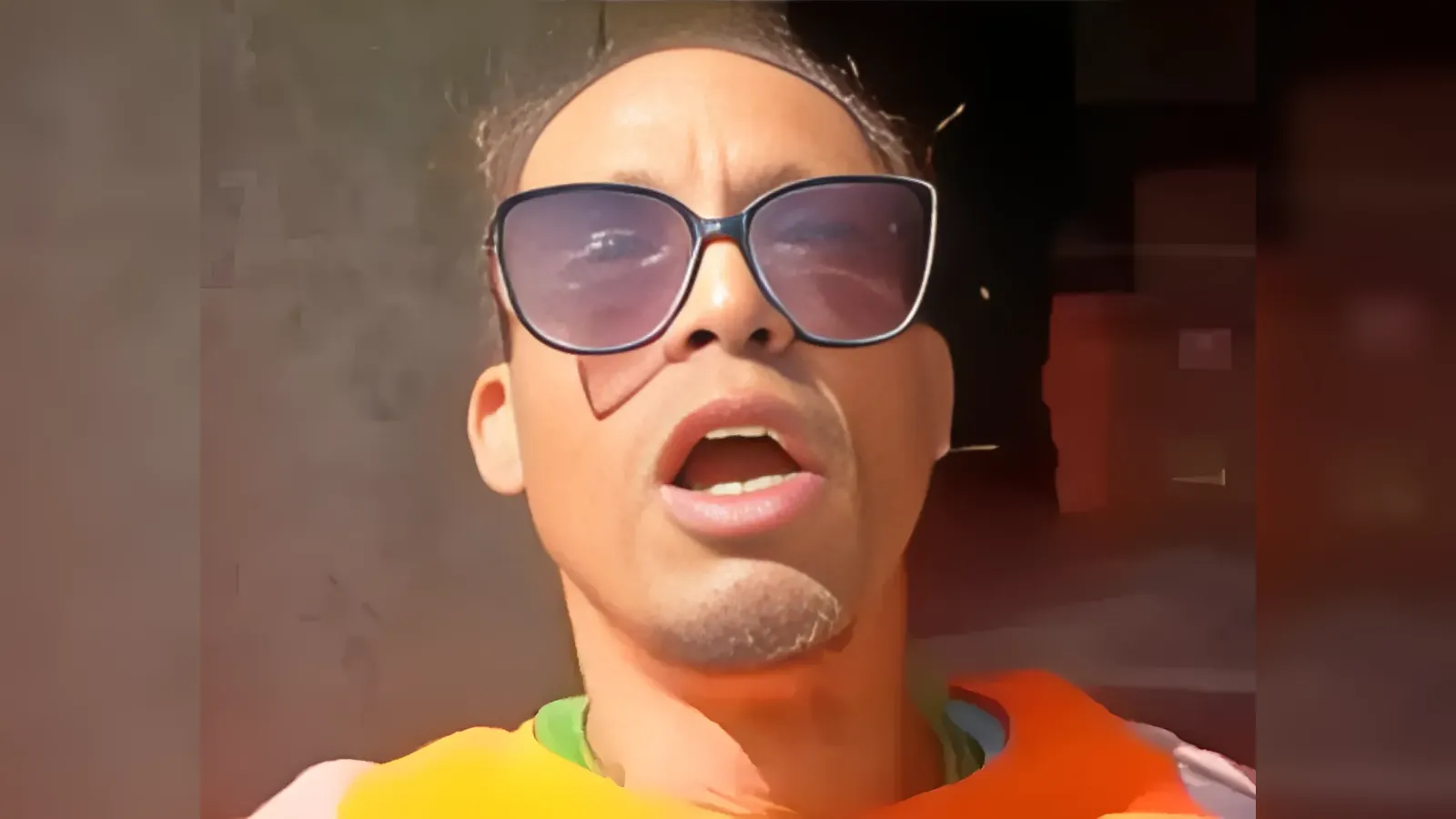 Close-up of a man wearing oversized sunglasses and an orange life jacket, his mouth open as if speaking or singing.