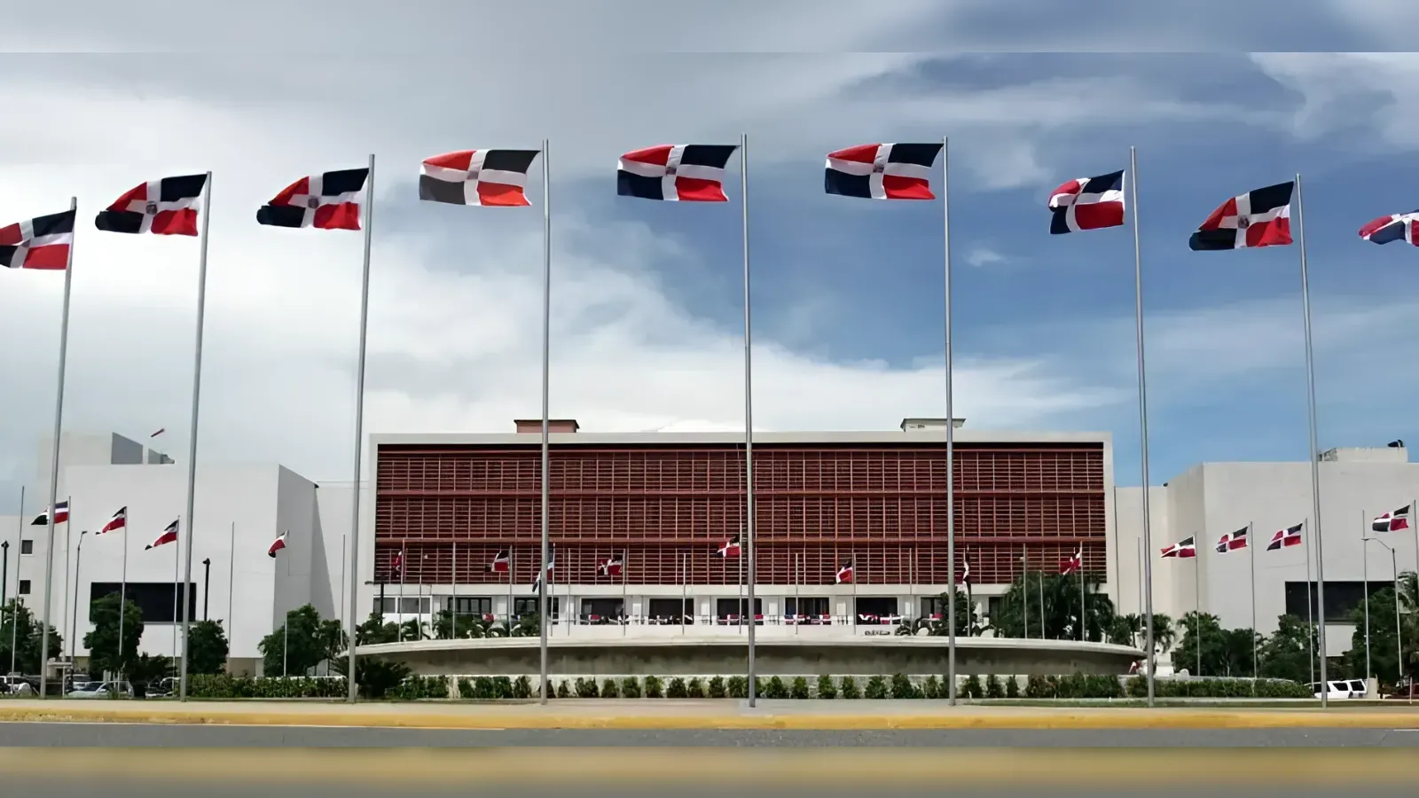Row of tall flagpoles with Dominican Republic flags in front of a modern white government-style building with red louvers and a circular-year-grade landscape.