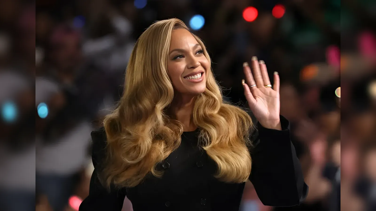 Smiling woman with long blonde hair waving to a crowd at an event in a black outfit.