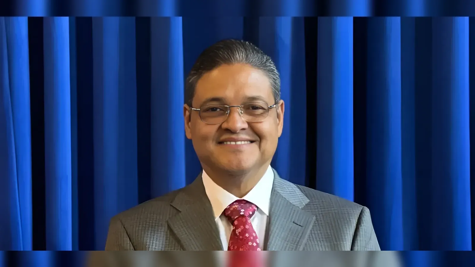 Smiling man in a gray suit and red/pink tie stands in front of blue stage curtains behind him.
