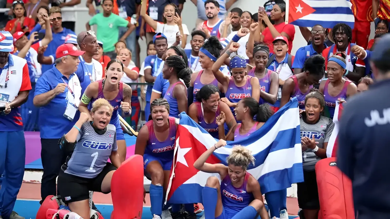Cuban women athletes in blue and purple uniforms celebrate a victory, waving a Cuban flag amid a cheering crowd.