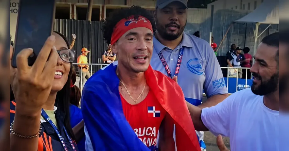 Man wearing a red bandana and Dominican flag draped over shoulders posing for a selfie with supporters at a sports event outdoors.