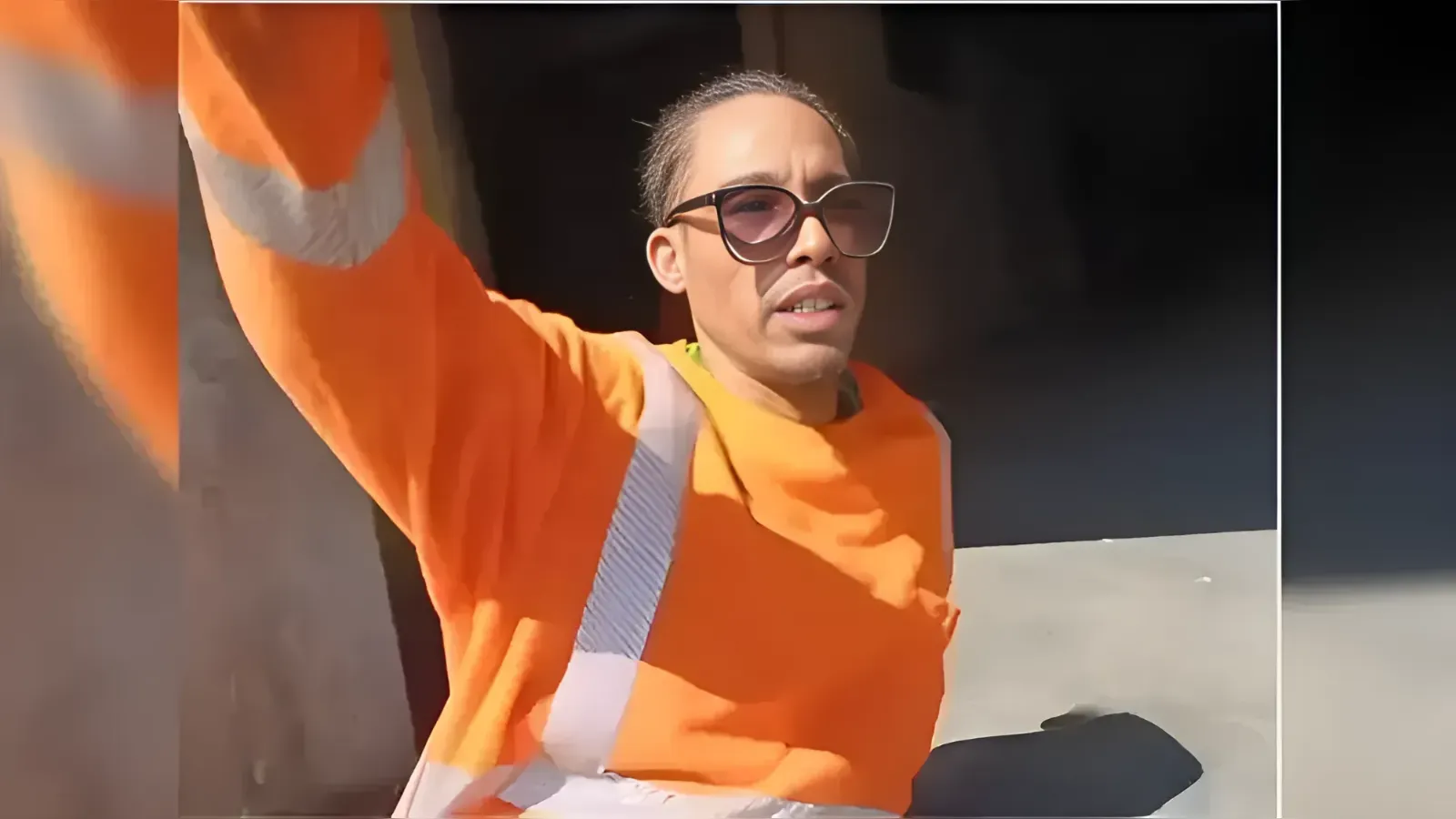 Person in a bright orange safety vest and sunglasses waves at the camera, outdoors.