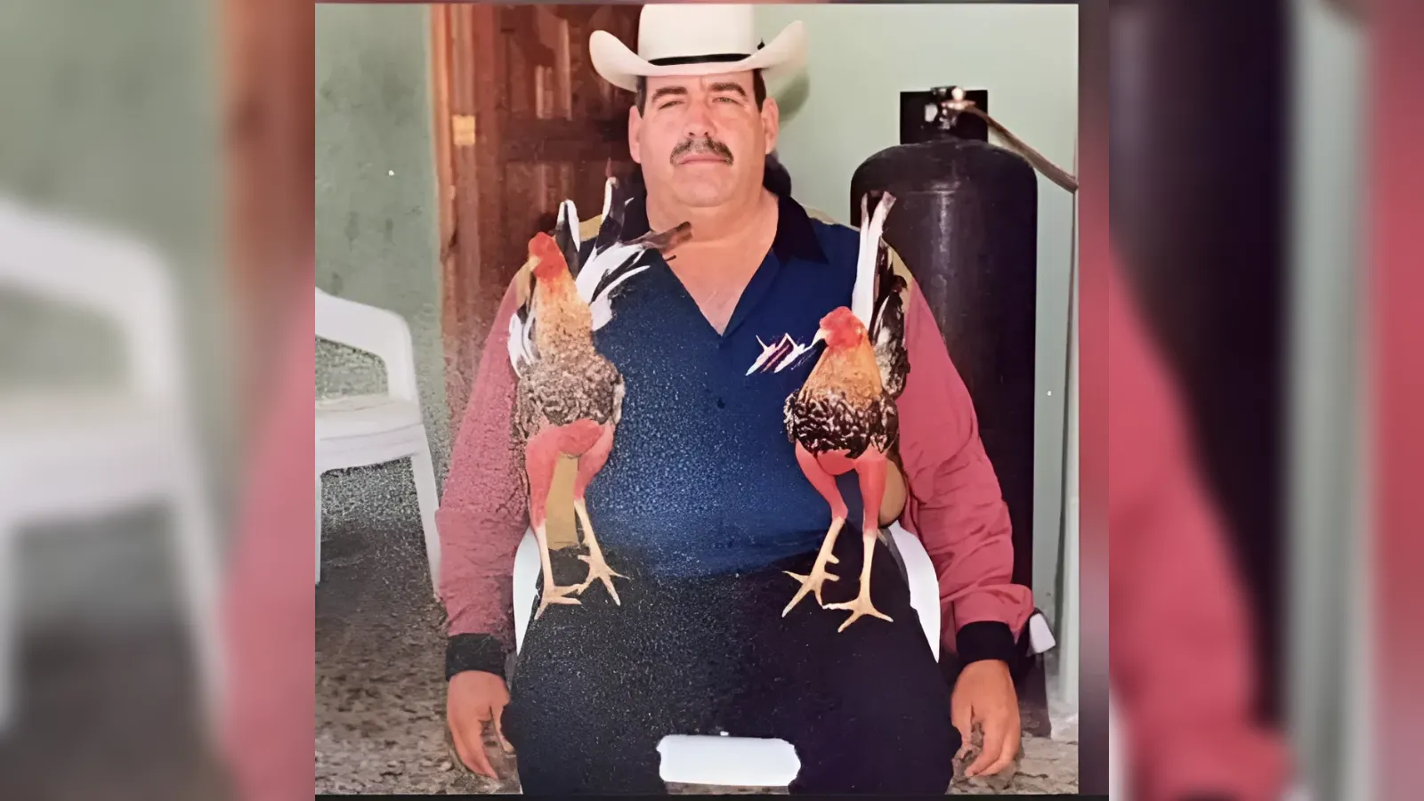 Man in a white cowboy hat and blue shirt sits with two roosters perched on his chest