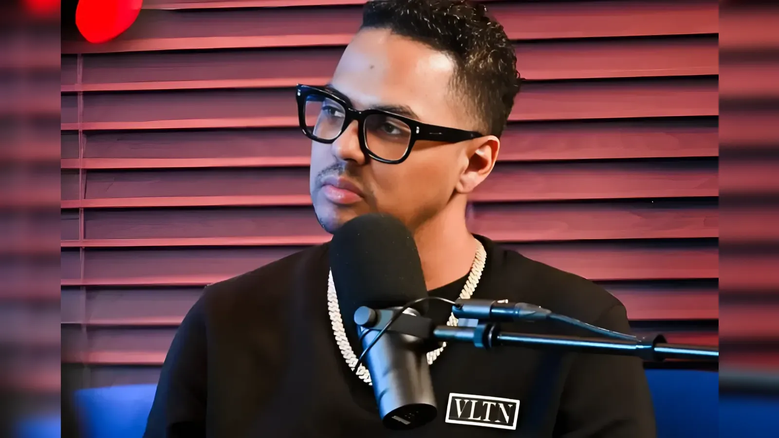 Man wearing black glasses and a chunky chain necklace speaks into a microphone in a recording studio with red blinds behind him.