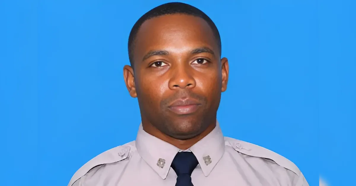 Portrait of a man in a light gray uniform shirt and navy tie against a solid blue background.
