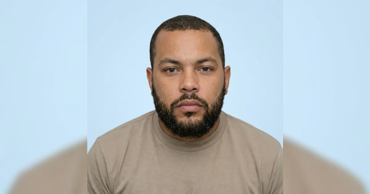 Portrait of a man with short dark hair and a full beard wearing a beige shirt against a light blue background.